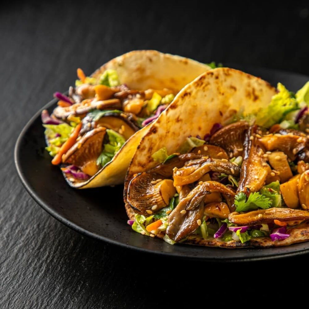 Two tacos with mushroom and vegetable filling on a black plate against a dark background
